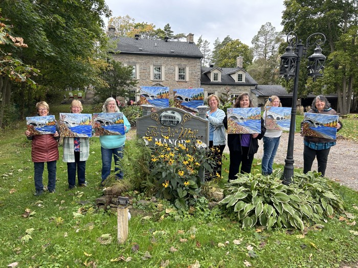 seven women holding paintings of pakenham bridge in front of stone mansion
