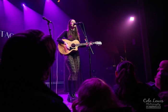 young woman singing and playing acoustic guitar on a stage lit with purple light