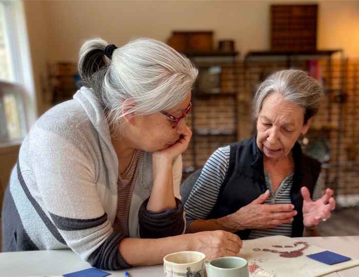 two women sitting at a table in an art studio, discussing a multimedia art project in front of them