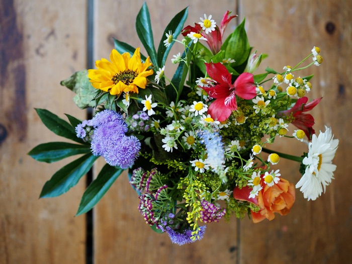 colorful mixed flower bouquet, including sunflowers, daisies, red lilies, orange roses, and purple statice, on a wooden surface