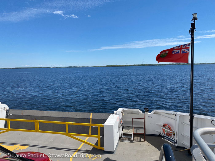 ontario flag flapping on deck of ferry with amherst island with wind turbines in distance