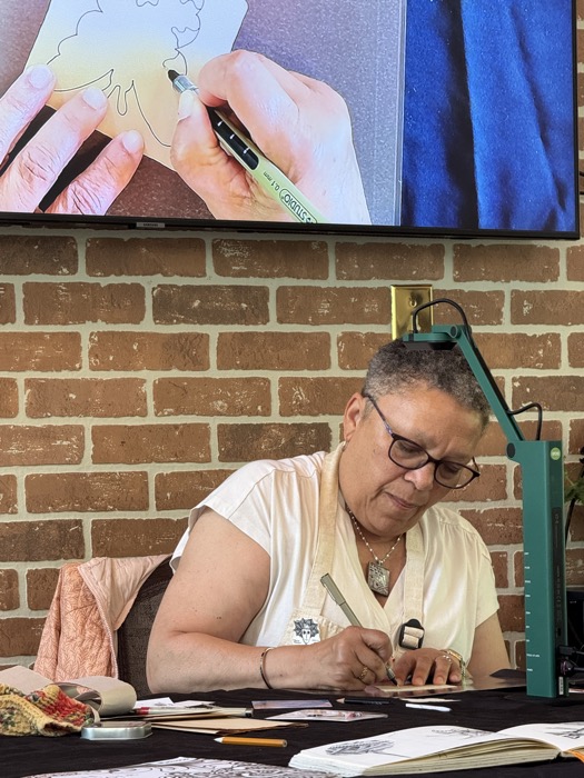 woman sitting at a table using marker to draw image on paper, which is projected on a screen above her head