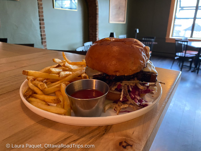 large sandwich on a brioche bun with fries on a white plate on a wooden bar