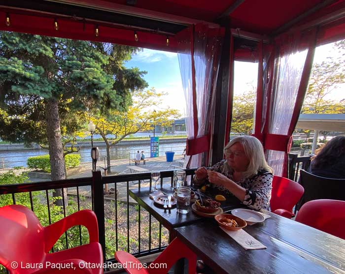 woman sitting at wooden restaurant table under an awning overlooking a river