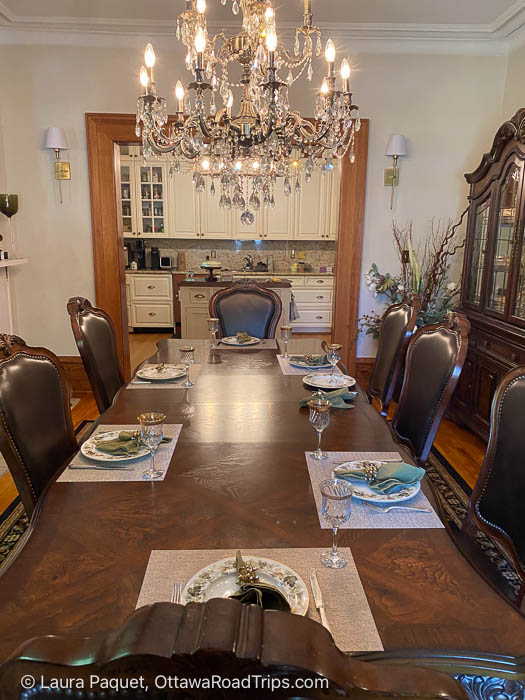 long antique table set with antique dishes, beneath a large chandelier, with kitchen in background