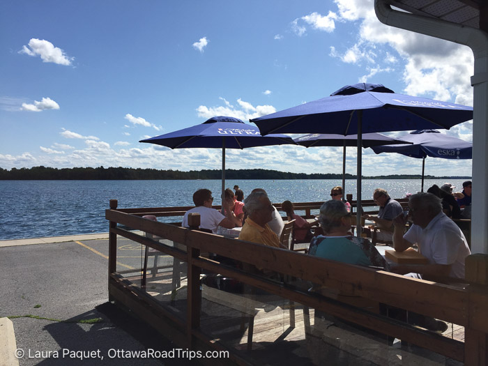 people sitting on a restaurant deck under blue patio umbrellas beside a river