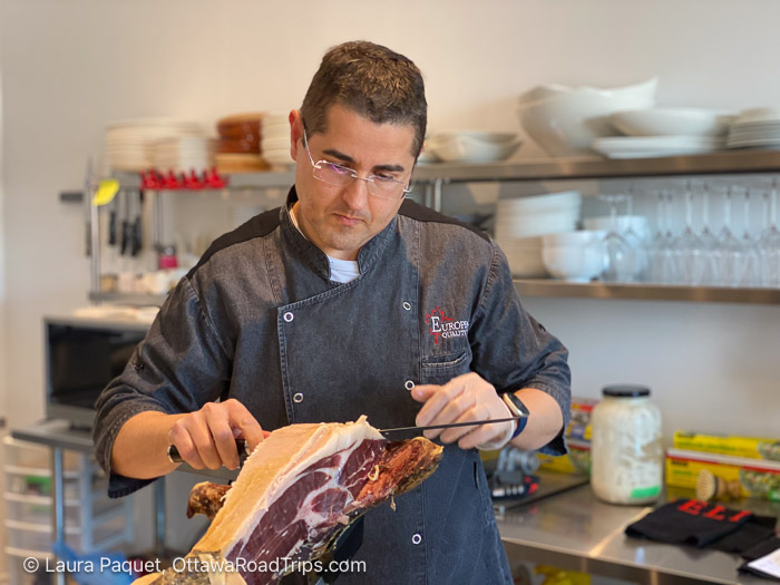 chef in a gray jacket carves a large cured ham on a cutting board in a bright kitchen setting.