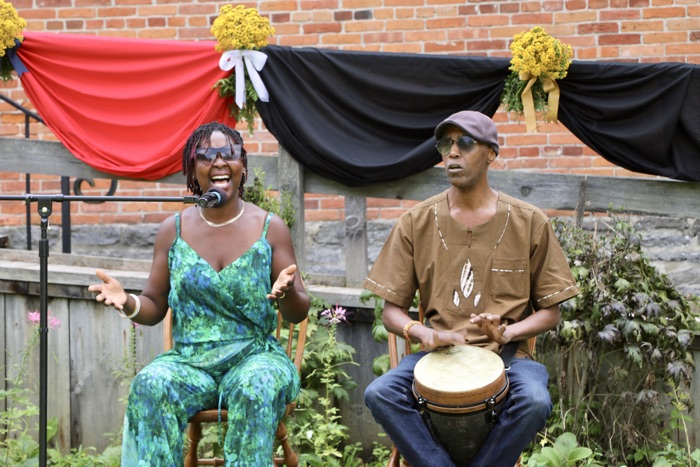 two musicians perform outdoors: a woman sings into a microphone while a man plays a drum beside her, with red and black draped fabric backdrop on a brick wall.