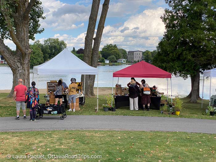 people browsing at outdoor stalls covered with awnings with pond and mill in background