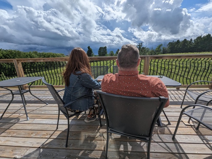 two people sit with their backs to the camera on a wooden deck, facing a green field and a cloudy sky.