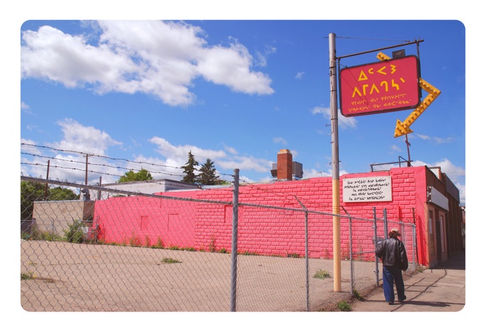 joi-arcand-ambermotors-hereonfutureearth - Ottawa Road Trips artwork of a pink cinder-block building with a sign in cree, next to a chain-link fence topped with barbed wire
