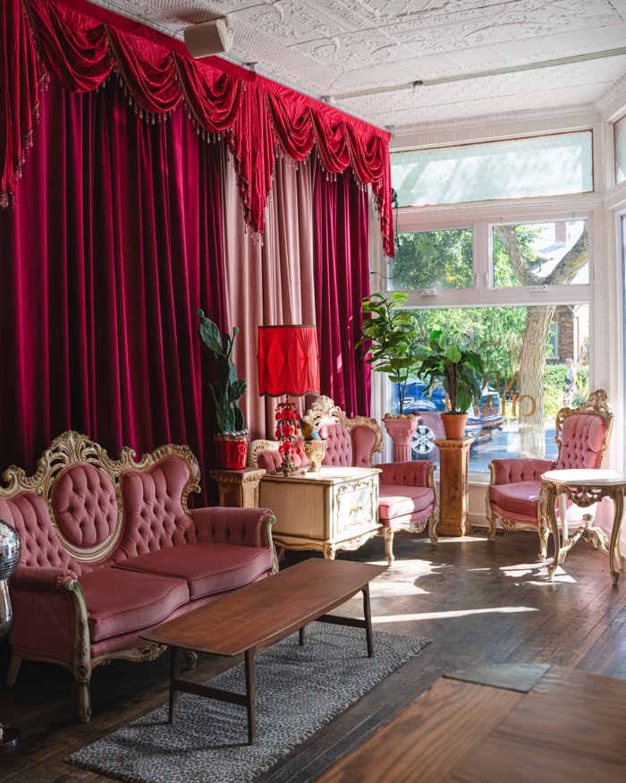 the neon chapel pec - Ottawa Road Trips vintage pink upholstered sofas and chairs in a sunlit lounge with burgundy velvet curtains, ornate gold furniture, and potted plants.